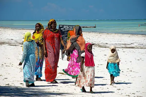 Kobiety i dzieci w kolorowych strojach spacerują po plaży Zanzibaru. Przed wyjazdem warto sprawdzić zalecenia dotyczące zanzibar szczepienia.