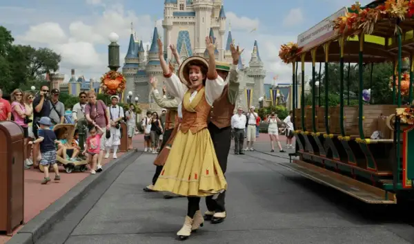 Uczestnicy parady w Disneylandzie pozdrawiają tłum. W tle zamek, a obok zabytkowy tramwaj.