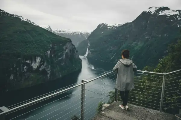 Kobieta podziwia majestatyczny fiord z wodospadem i ośnieżonymi szczytami w tle.