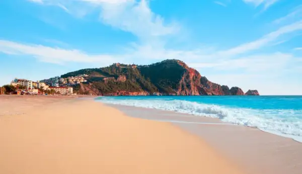 Plaża Kleopatry w Alanyi, z turkusowym morzem, złotym piaskiem i malowniczymi wzgórzami z zamkiem.