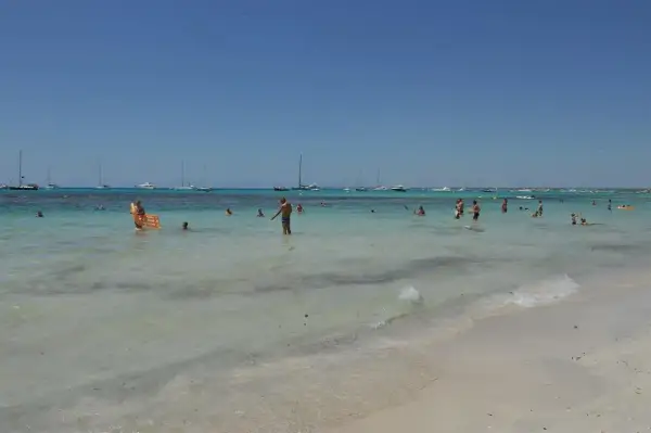 Słoneczny dzień na plaży Es Trenc. Ludzie kąpią się w turkusowej wodzie, na horyzoncie widać żaglówki.