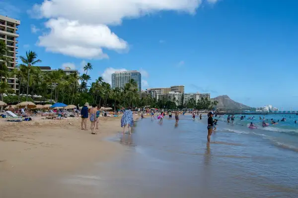 Słoneczny dzień na plaży Waikiki, z turystami kąpiącymi się w oceanie i odpoczywającymi na piasku, w tle palmy i hotele.