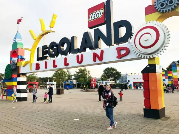 Uśmiechnięta dziewczyna z plecakiem przed wejściem do Legolandu. W tle widać bramę z napisem "LEGOLAND BILLUND" i budki z biletami.